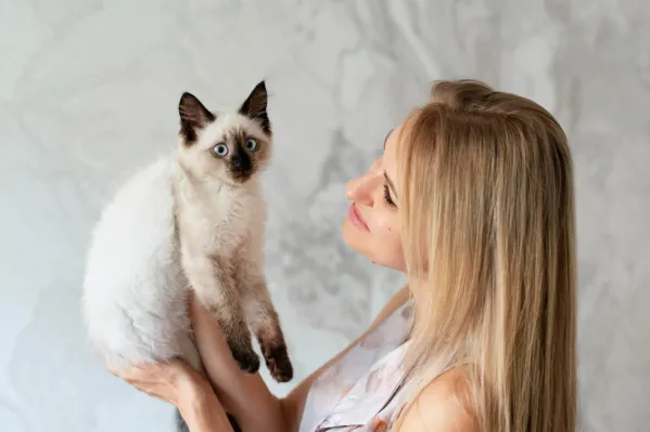 Blondýna drží v náručí siamské koťátko, které s vytřeštěným pohledem pozoruje okolí.