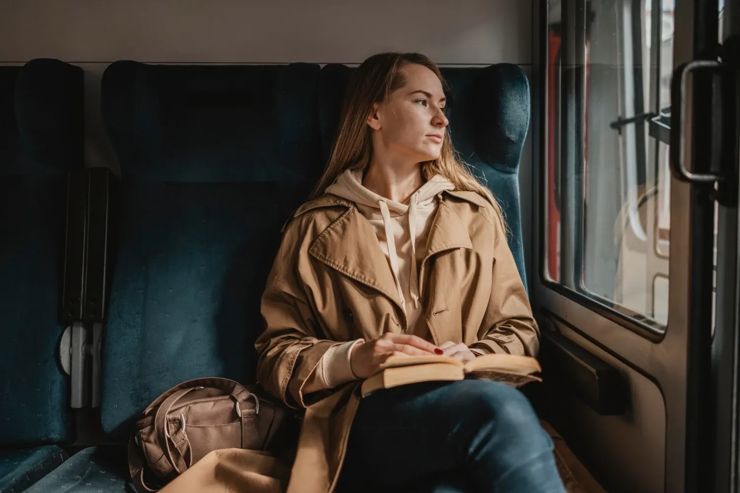 Žena s dlouhými vlasy v kabátu sedí ve vlaku u okna a čte.